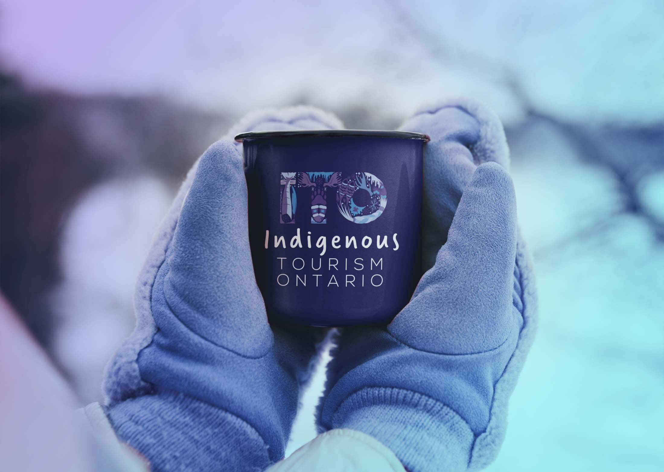 Two hands covered with light purple mittens hold a tin mug with the ITO - Indigenous Tourism Ontario logo
