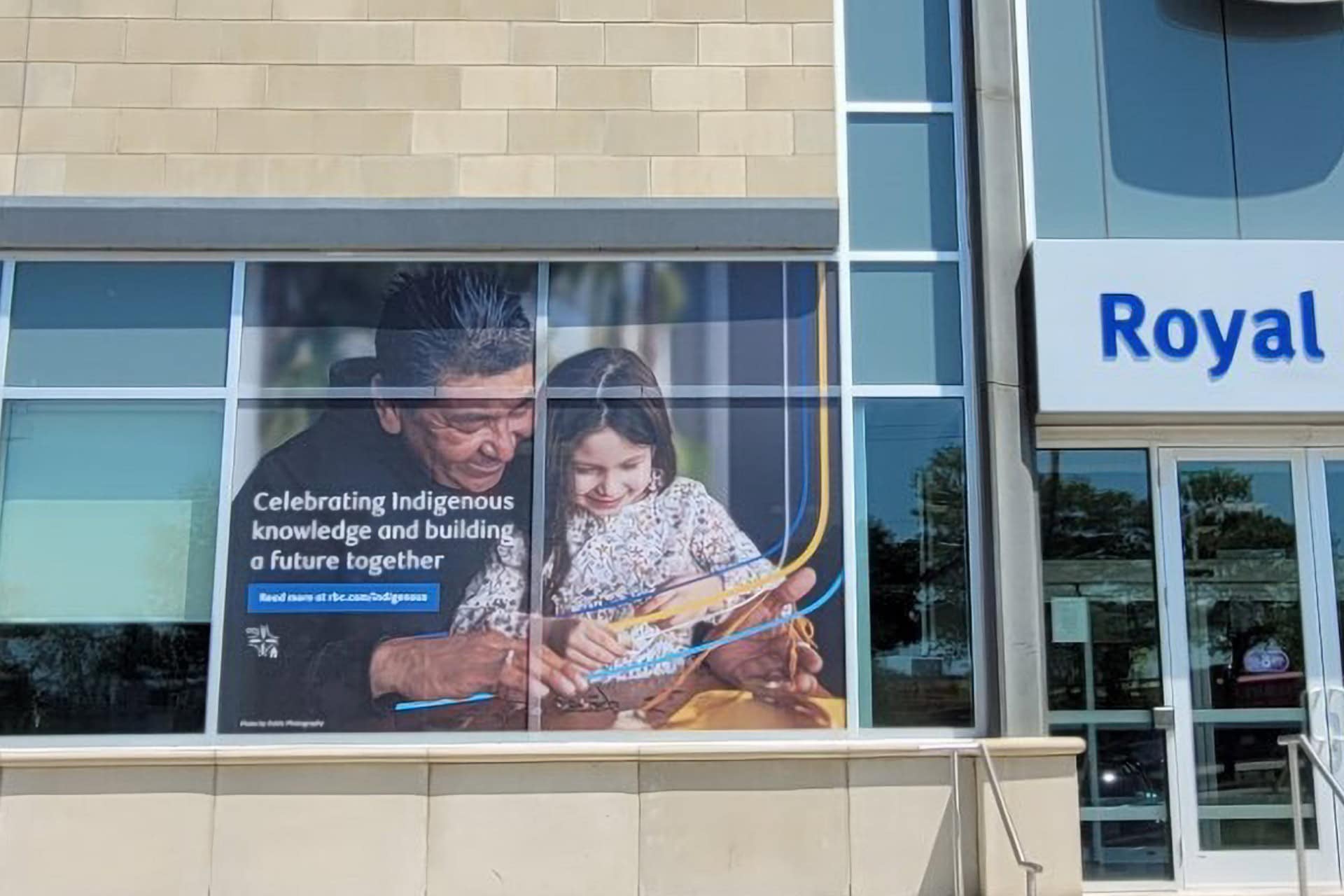 An exterior shot of an RBC branch. On the windows of the branch is a large design showing an Indigenous man and a young girl looking at something together. The text reads 'Celebrating Indigenous knowledge and building a future together' over top of a link to a webpage.