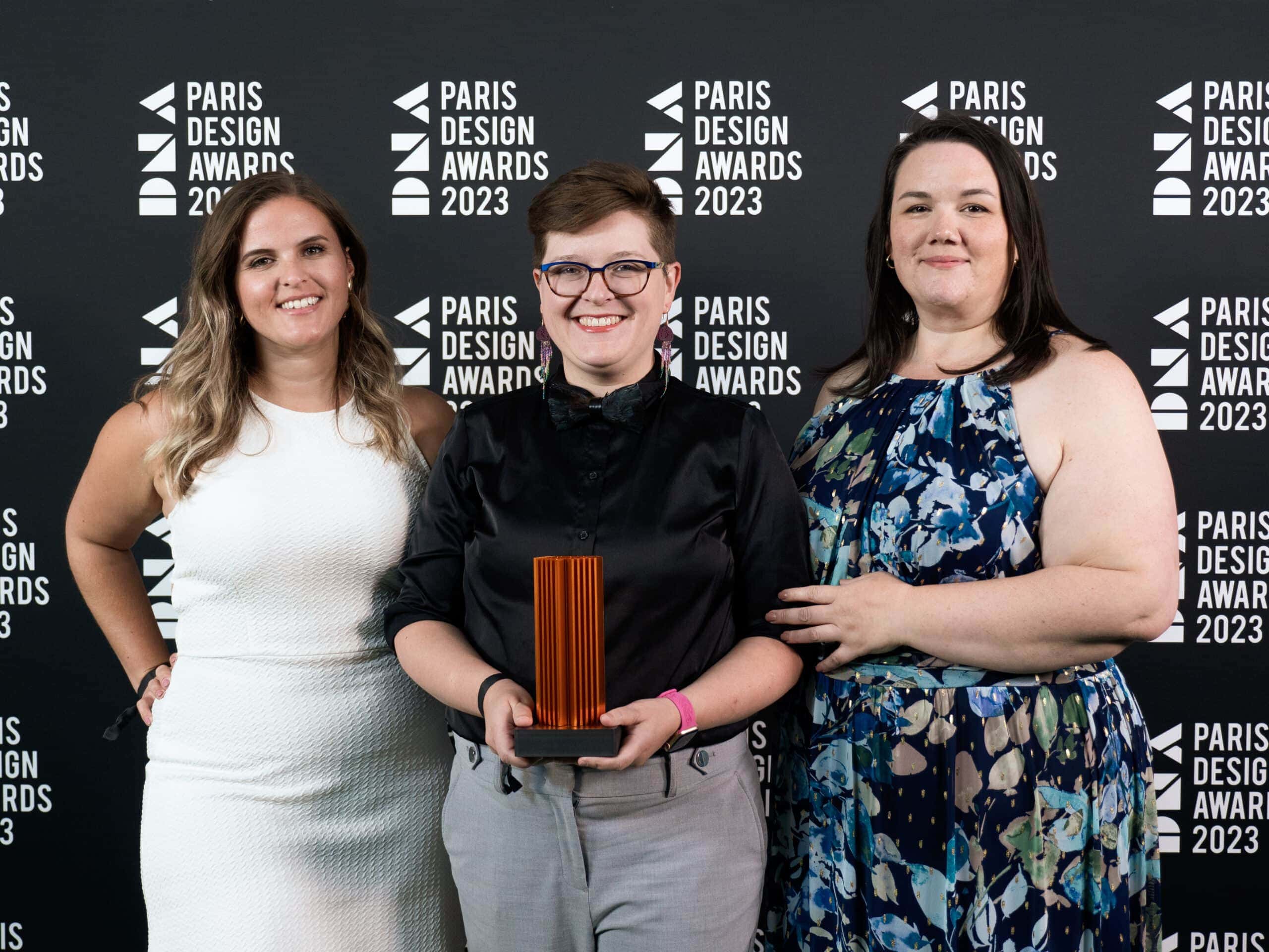 Jennica Robinson, Meggan Van Harten, and Jennifer Taback stand in front of a wall covered with the 'DNA Paris Design Awards 2023'. Meggan stands in the middle holding the award in her hands.