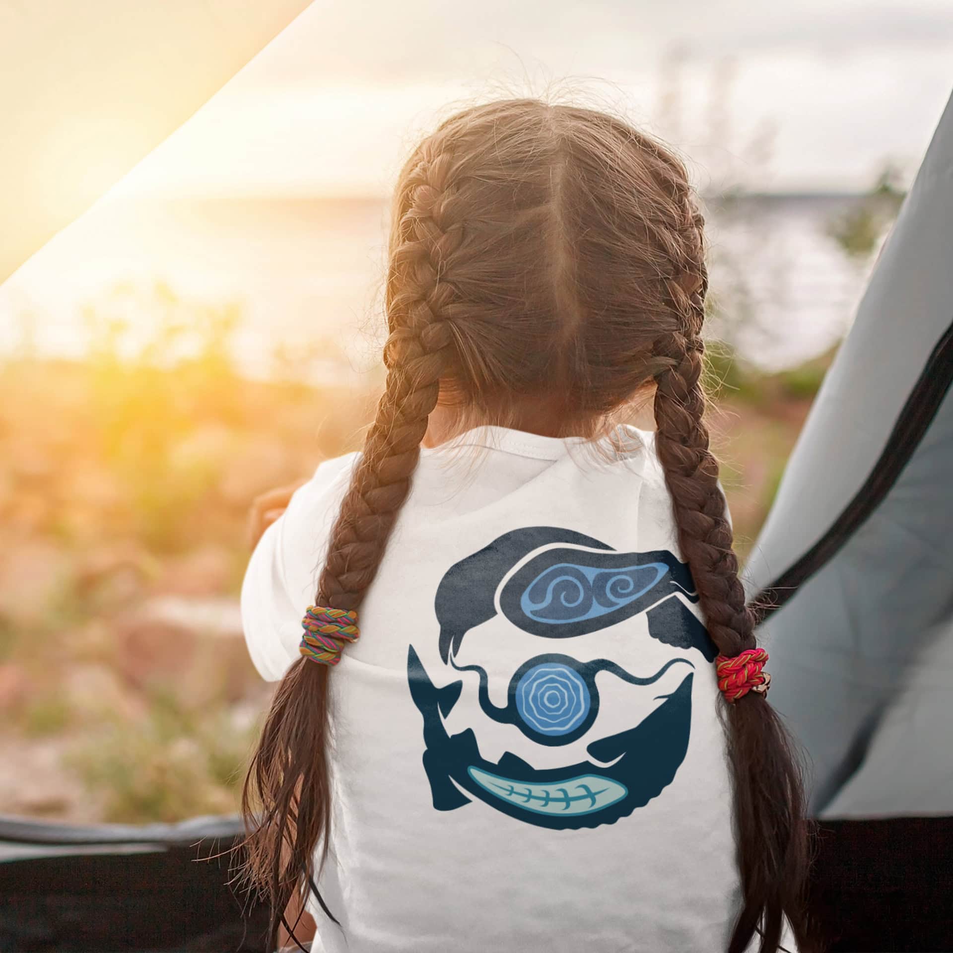 A young girl stands, looking out over the Great Lakes. On her white t-shirt is a large Biinaagami logo. Her braids fall down her back on either side of the logo.