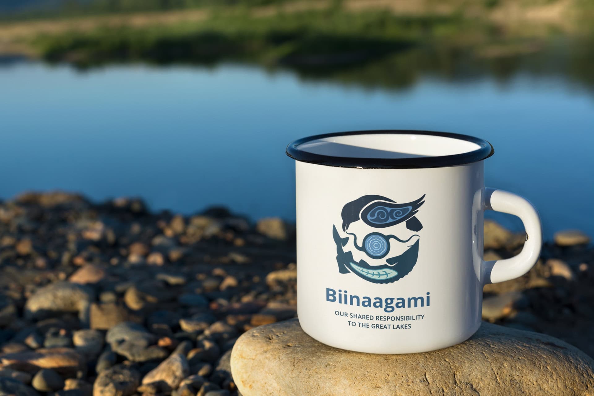 A cup with the Biinaagami logo sits next to the waters of the Great Lakes.