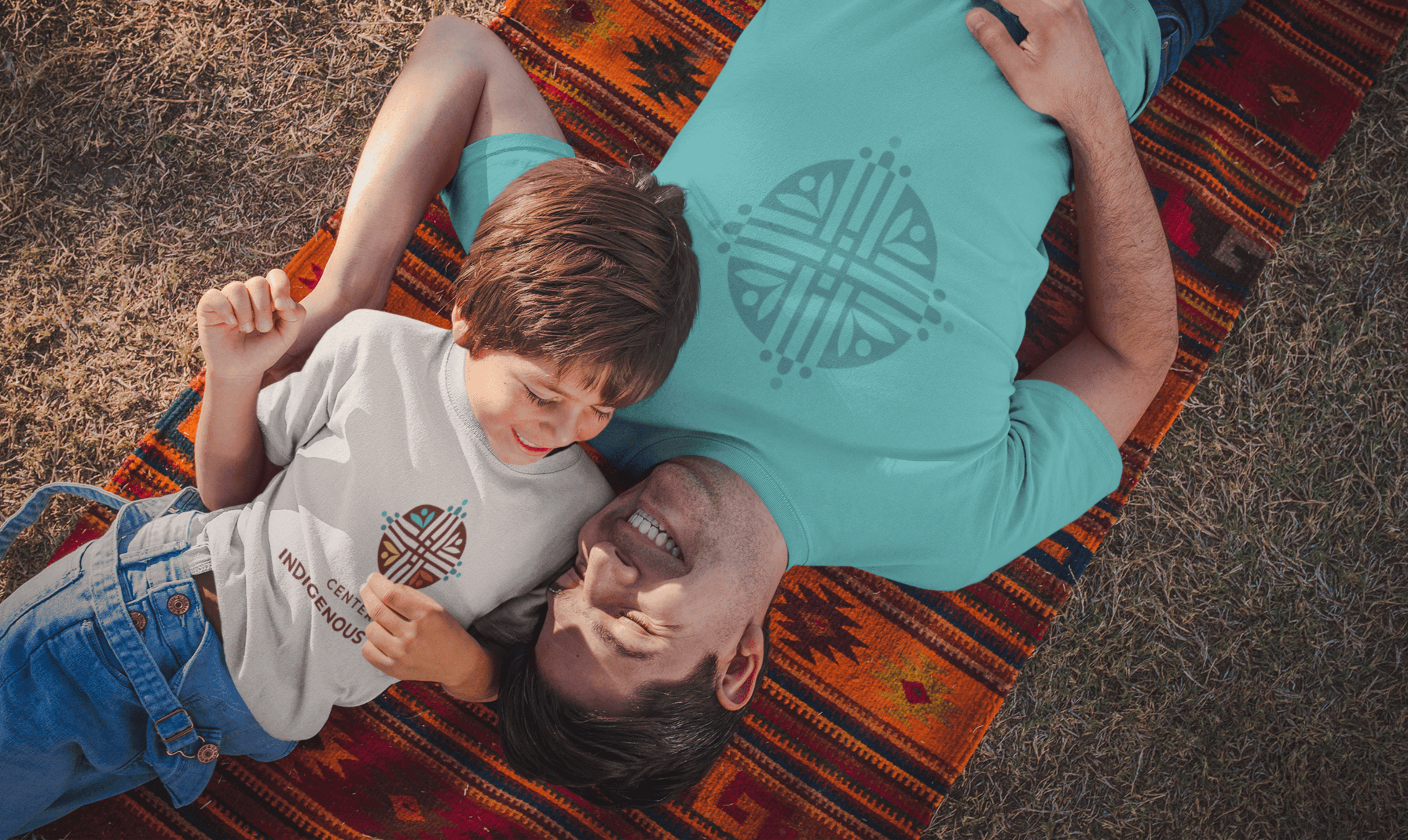 A father and son lay shoulder to shoulder on a blanket on grass. The man's t-shirt has a large logo of the Centre for Indigenous Health. The little boy also has a full colour version of the logo on his white t-shirt. They are smiling and laughing.