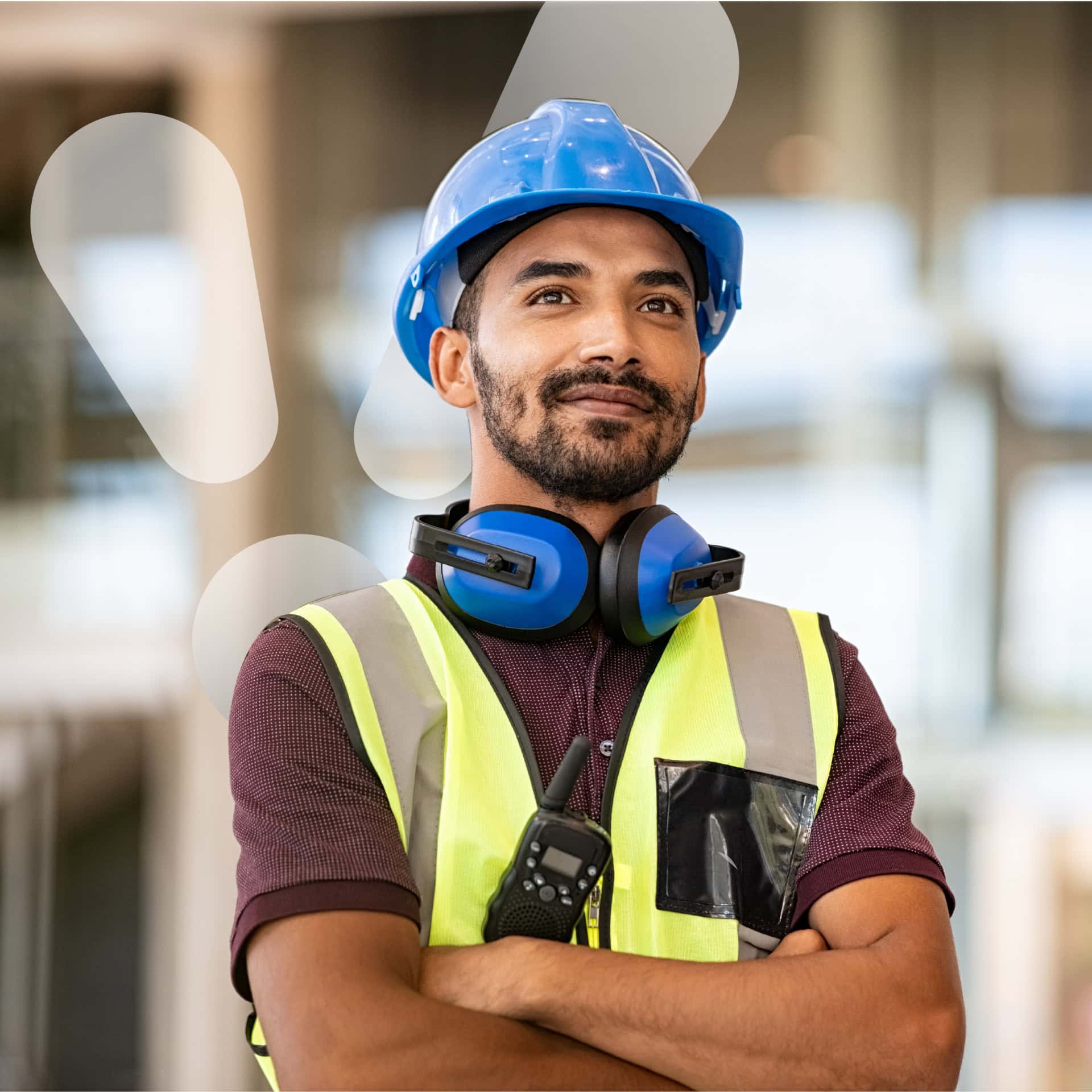 A man wearing a blue construction hat and ear protection stands proudly with arms crossed. He is wearing a high visibility vest for safety while working at a sustainable energy site.