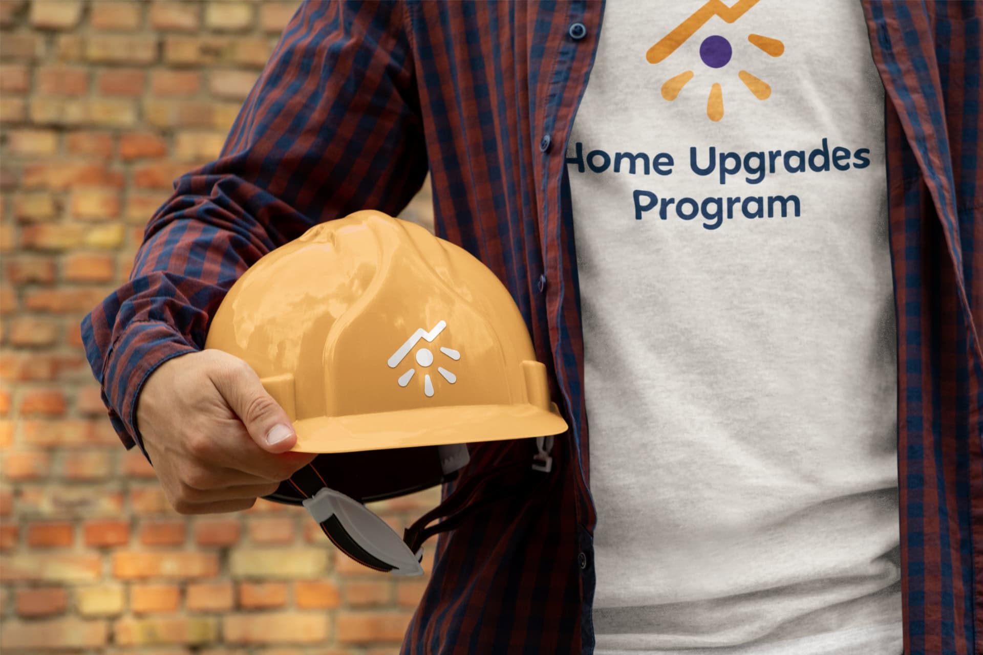 A man stands in front of a brick wall. We just see his chest and arm. Under his arm is a yellow construction hat. His shirt reads 'Home Upgrades Program' with the Kambo logo on it.
