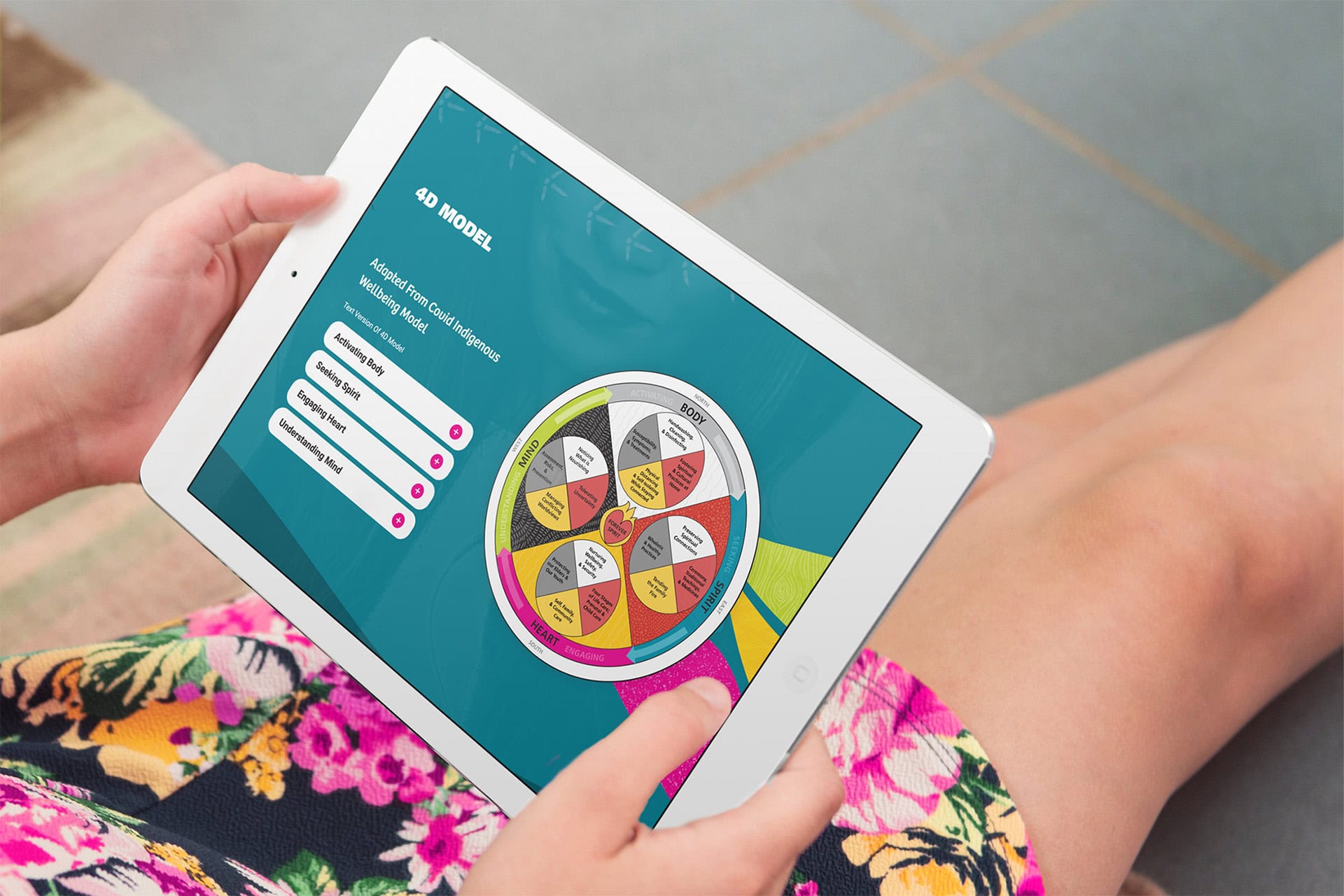 A woman in a floral dress holds a tablet in her hands. On it is a Centre for Wise Practices webpage showing a complex Medicine Wheel diagram.