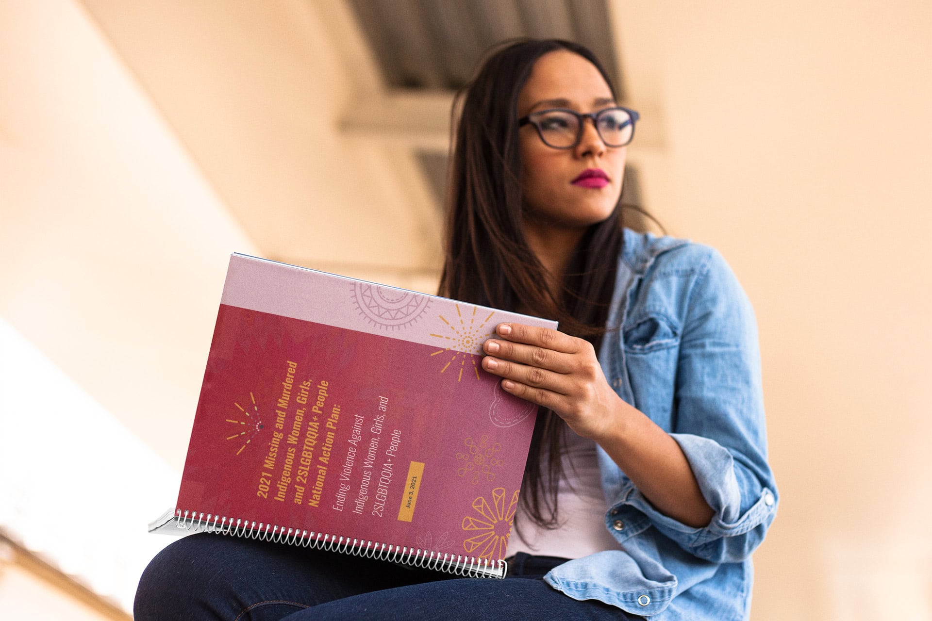 A young Indigenous woman in casual clothes and glasses sits holding a copy of the National Action Plan open on her knee. From the looks of the hallway she is in, she appears to be a college student.