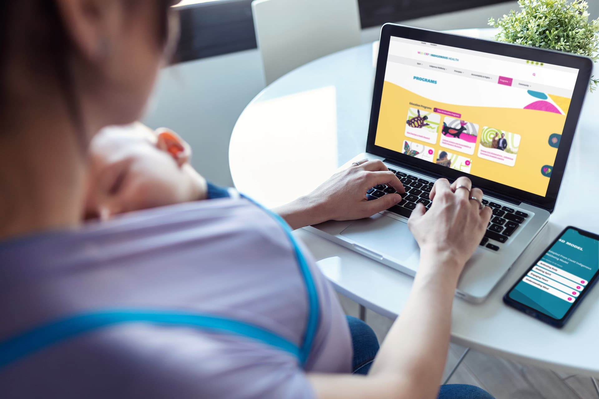 A young mother viewing the CWP website on her laptop. Her baby is asleep in a baby carrier tucked against her chest under her purple shirt.