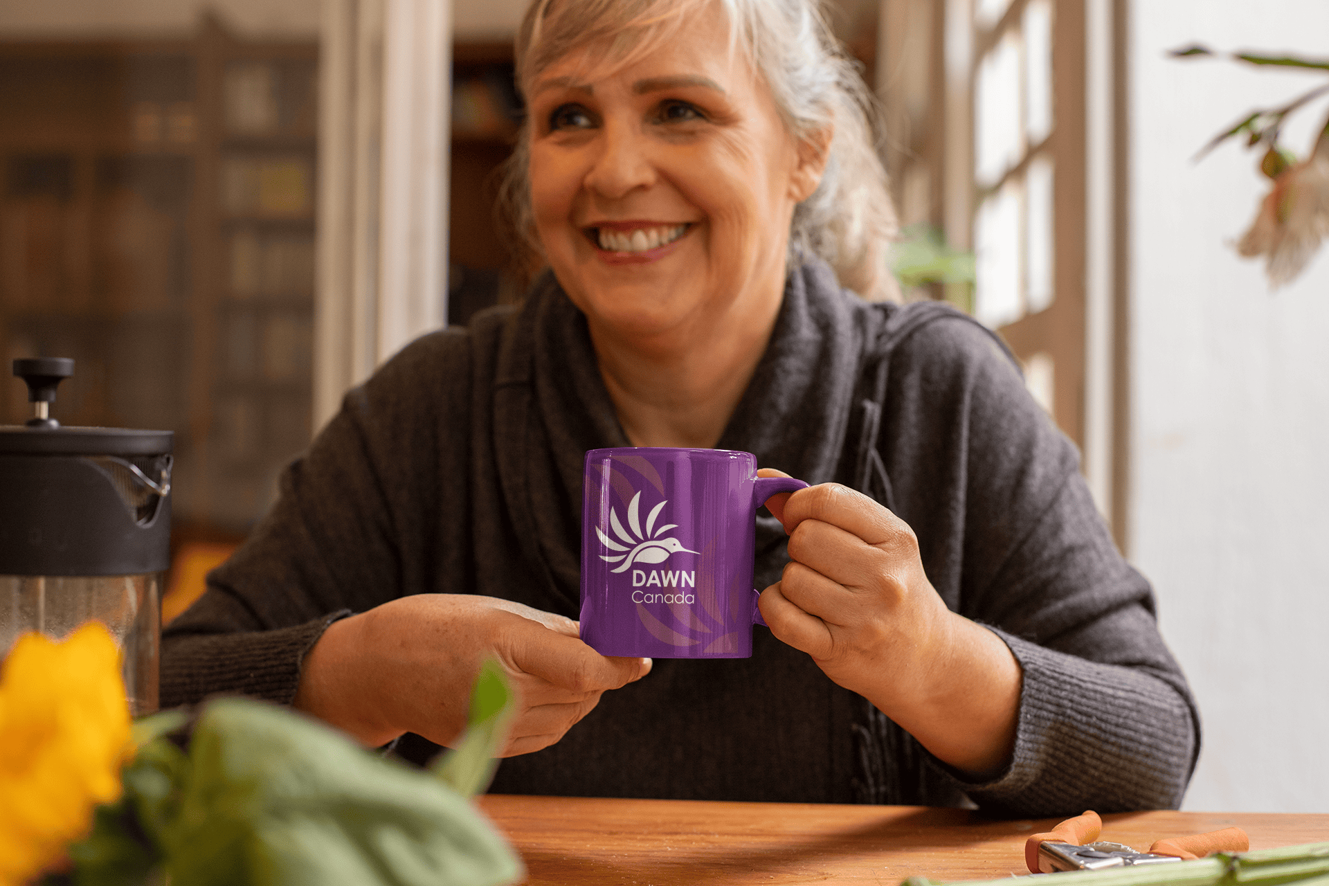 A warm smiling middle-aged woman in a sunny breakfast nook holding a purple mug with a white DAWN Canada Logo