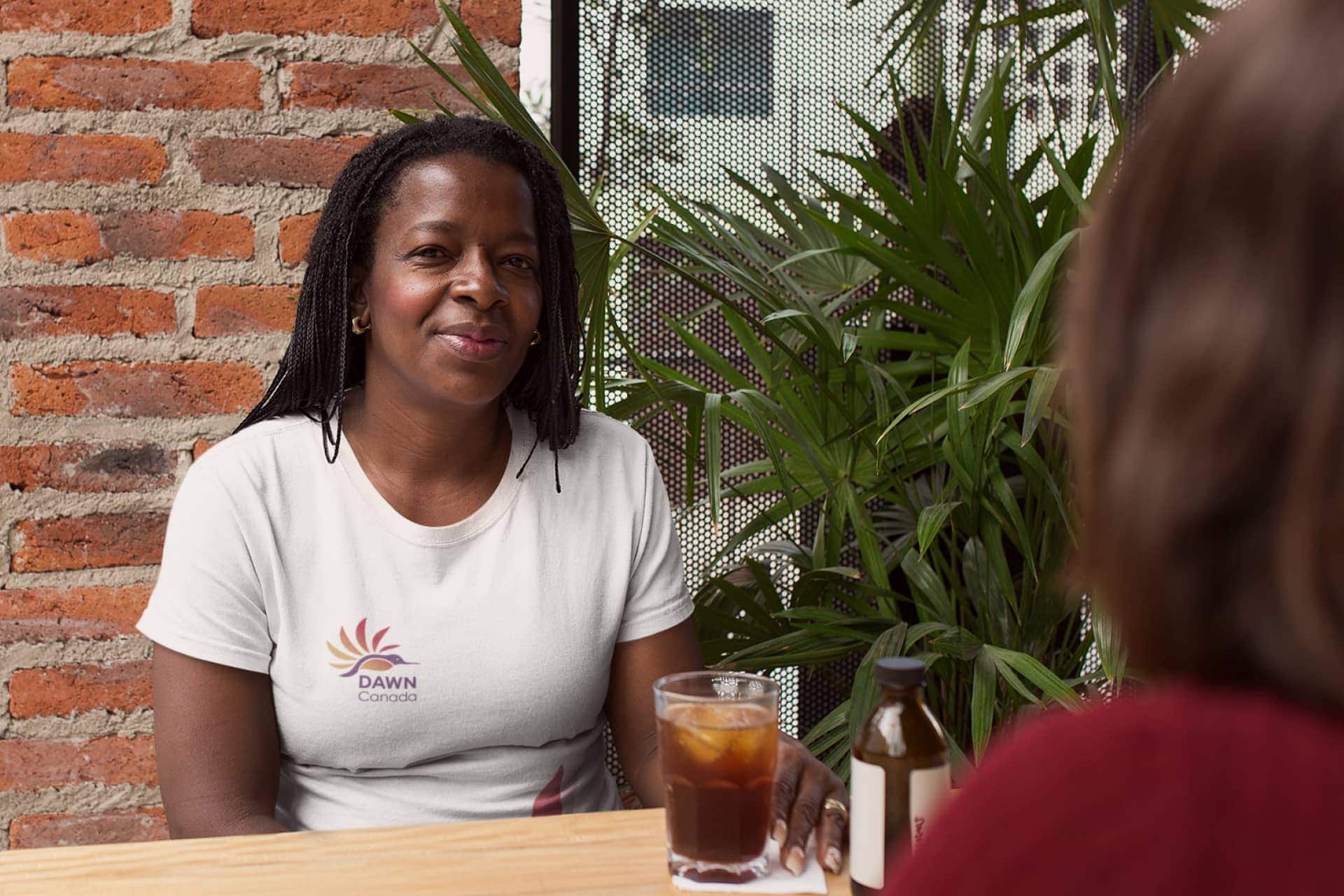 Middle-aged smiling Black woman having a tea, wearing a white t-shirt featuring the DAWN logo on the chest.