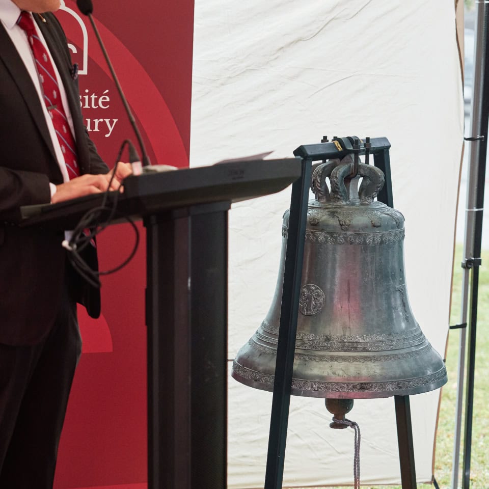 Person speaking at a podium in front of a Université de Sudbury banner, next to a bell.