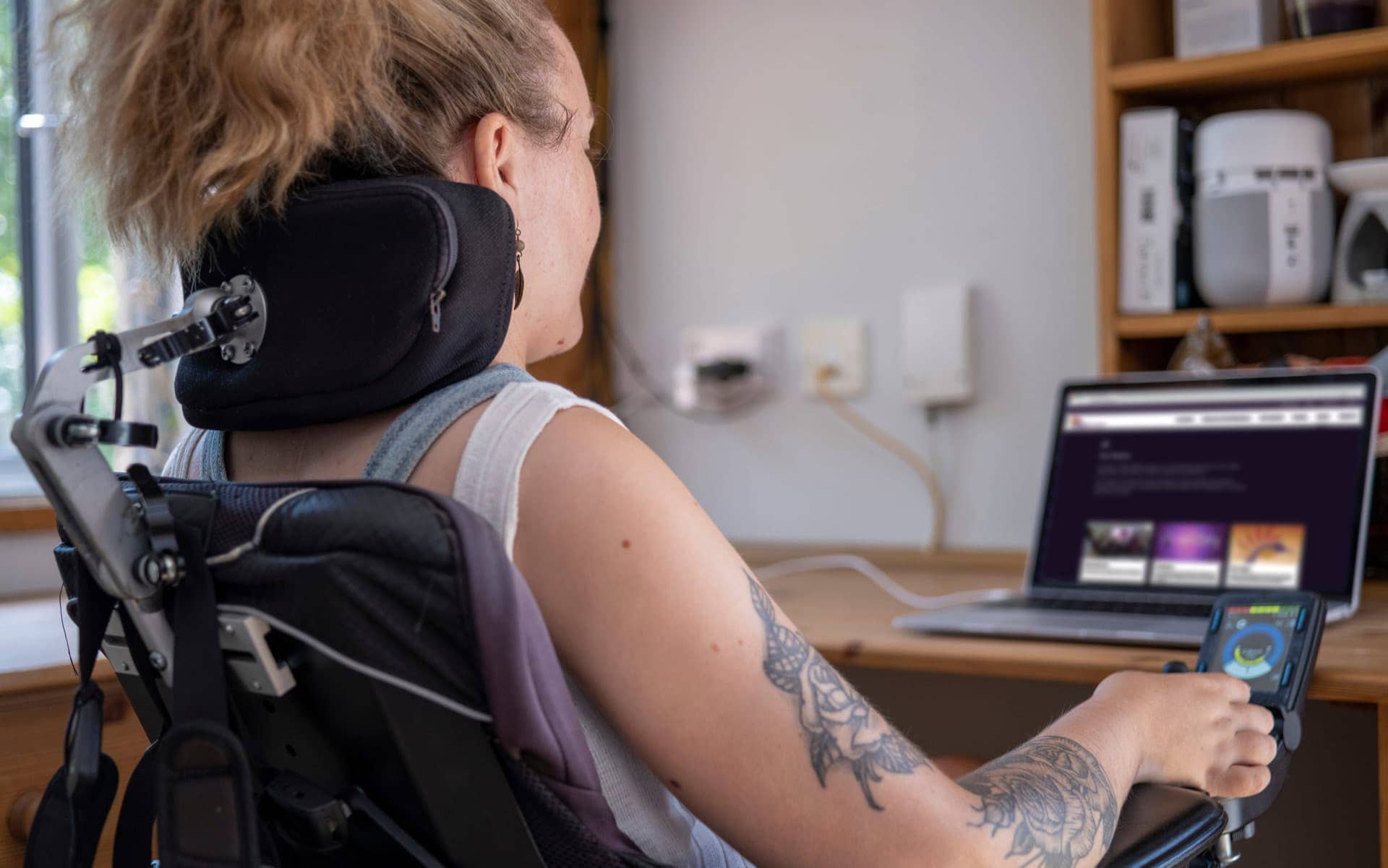 A young curly-haired woman in a wheelchair with neck support and hand controls looks at a laptop with the DAWN Canada website.