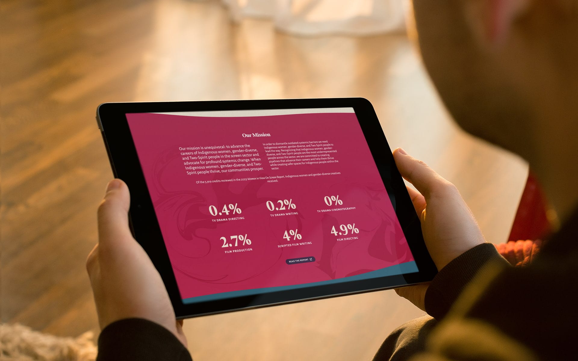 Someone holding a tablet in both hands showing the Mission as it's listed on the website, including the following statistics. Of the 5,919 credits reviewed in the 2023 Women in View On Screen Report, Indigenous women and gender-diverse creatives received: 0.4% TV drama directing, 0.2% TV drama writing, 0% TV drama cinematography, 2.7% film production, 4% scripted film writing, 4.9% film directing.