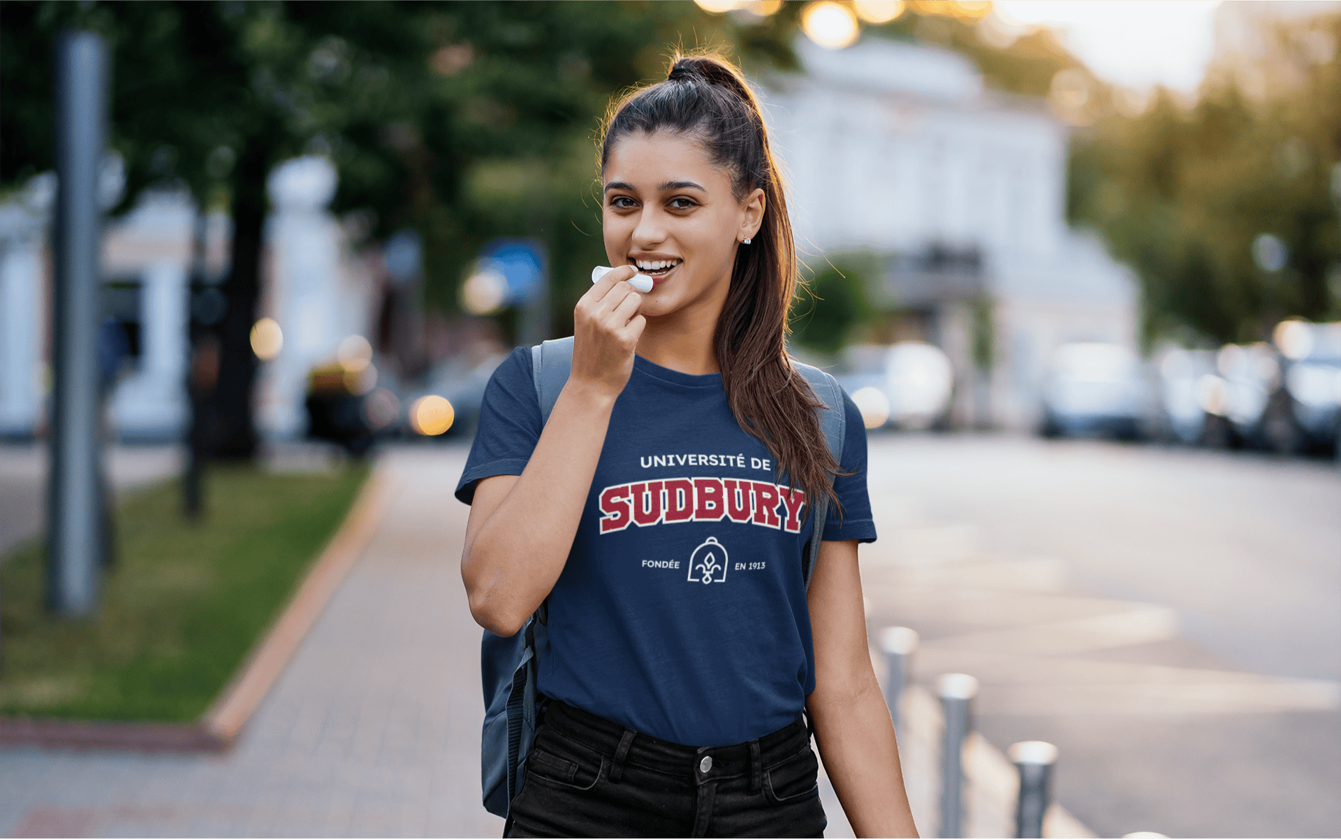 Student standing outside near the road with a high pony tail, applying lip gloss and smiling. She is wearing a navy blue Université de Sudbury tshirt