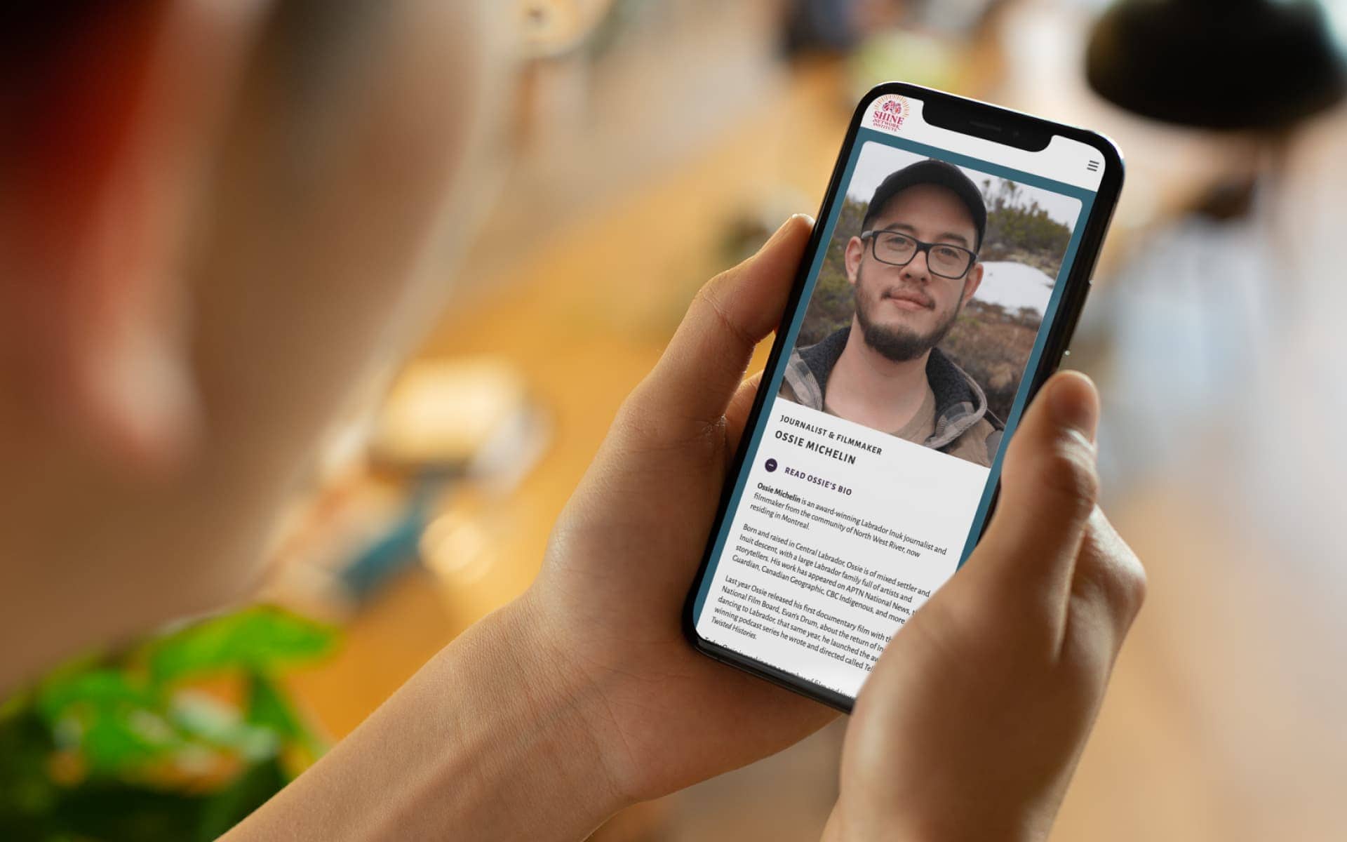 Someone holding a phone displaying a profile on the SHINE Network Institute website with details about the trans-gender award-winning Labrador Inuk journalist and filmmaker, Ossie Michelin.