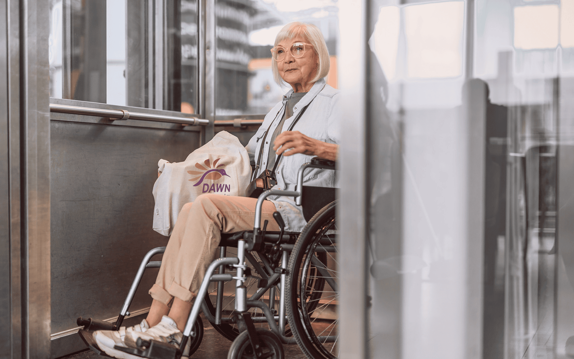 An elderly urban woman in a wheelchair, with shoulder length white hair and pink glasses exits an upscale elevator while holding a large tote bag with part of the DAWN Canada logo showing.