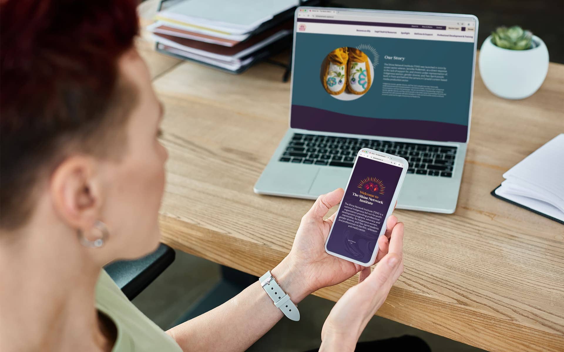 Short-haired person with earrings in a comfortable office, holding a cellphone with the SHINE Network Institute website featured.