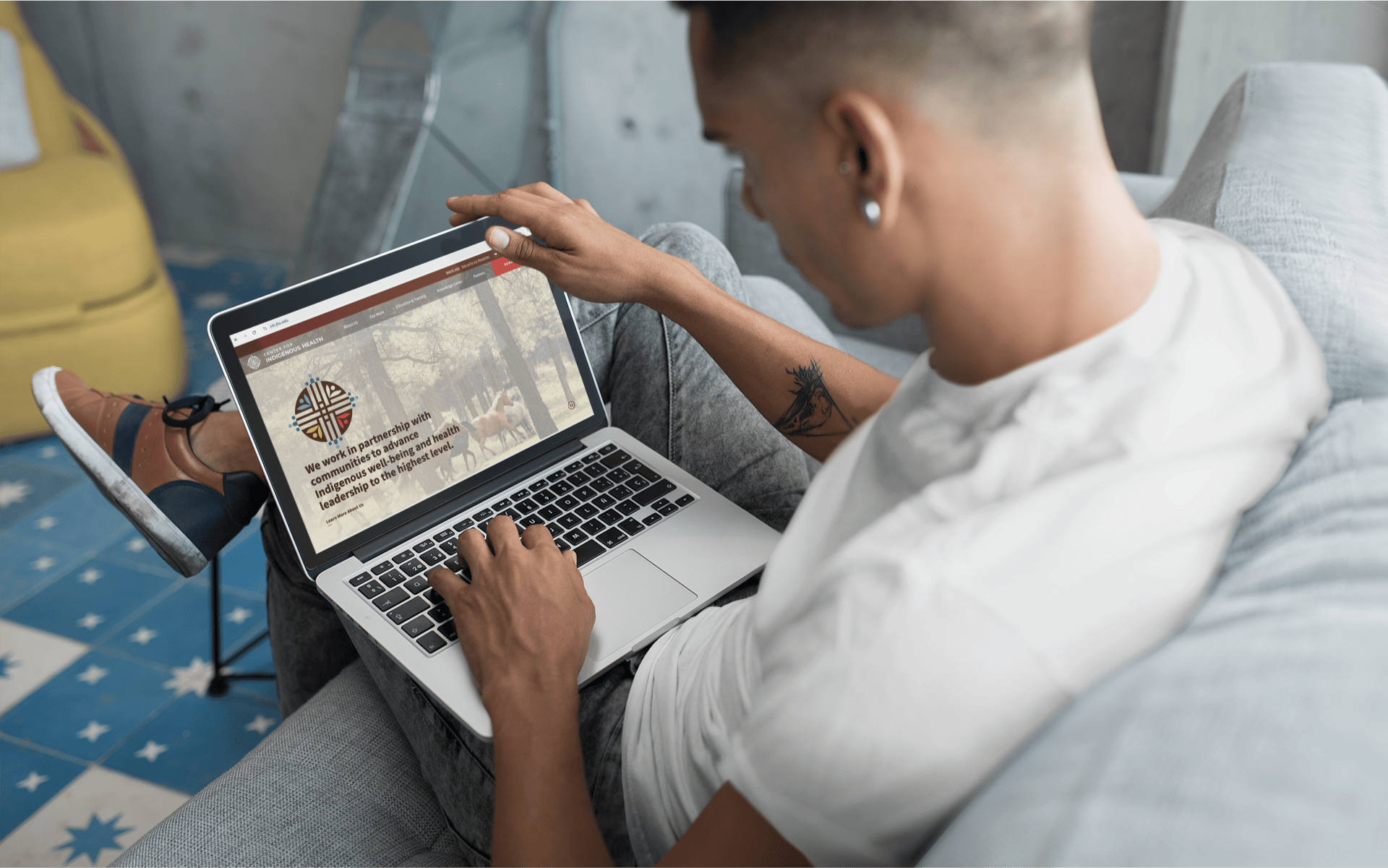 You man lounging on a couch with a laptop in his lap displaying the home page of the website for the Center of Indigenous Health