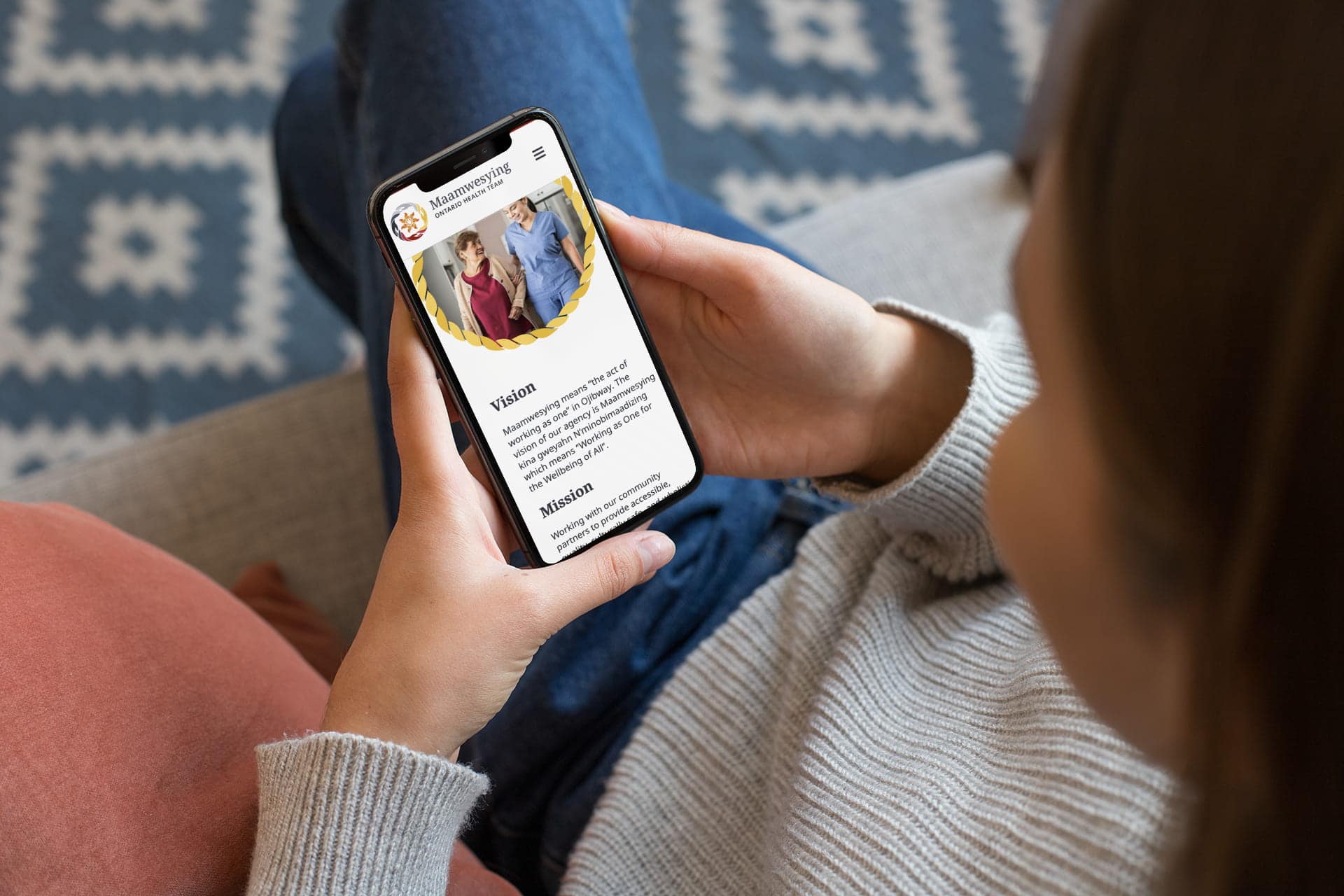 A young adult holding a cell phone comfortably at home. The cell phone shows the Maamwesying website open to a section describing the Vision, which reads "Maamwesying means "the act of working as one" in Ojibway. The vision of our agency is Maamwesying kina gweyahn N'minobimaadizing which means "Working as One for the Wellbeing of All"."