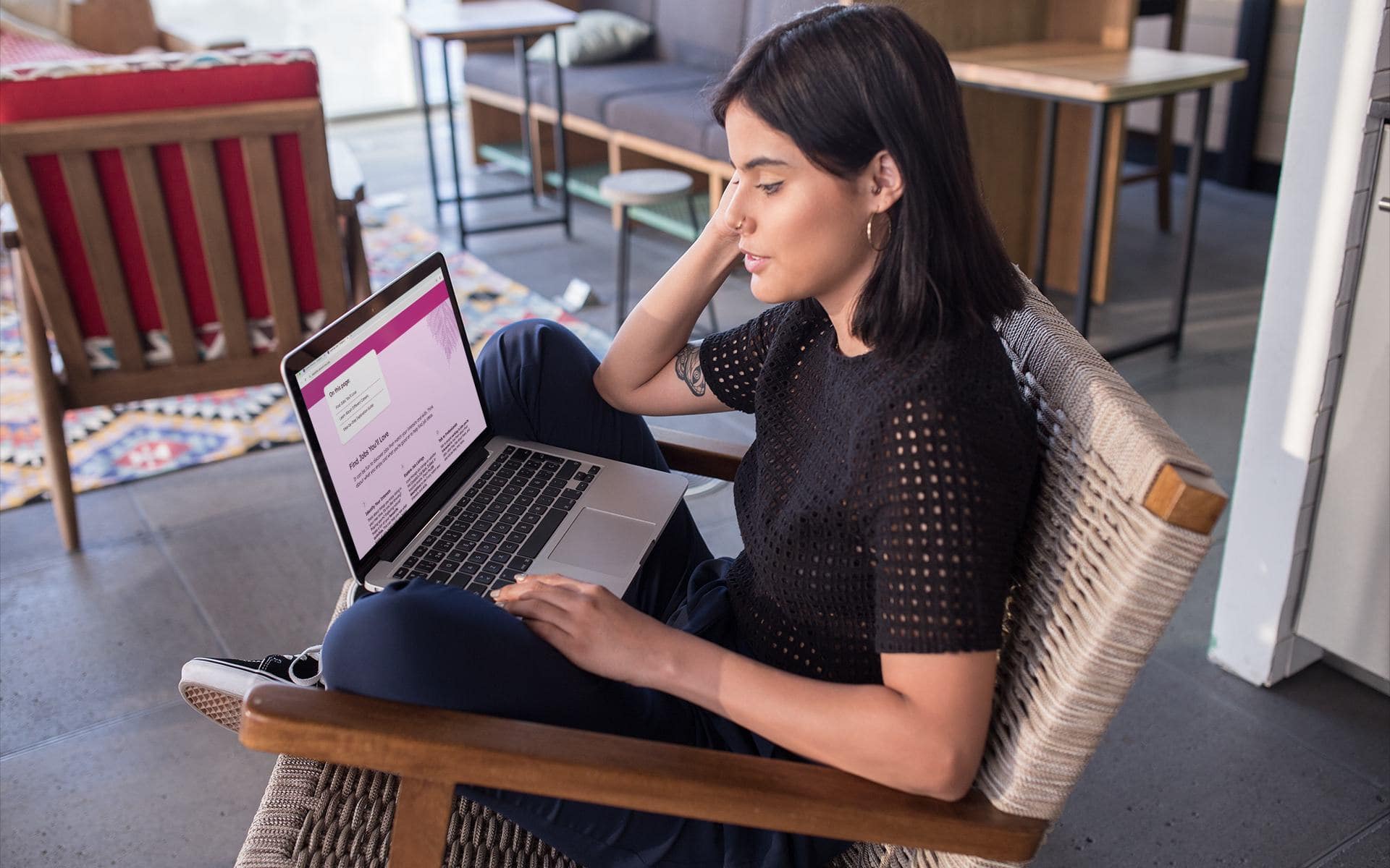 Young woman with shoulder-length dark hair sitting cross-legged on an accent chair in a living room, using a laptop to access the Congress of Aboriginal Peoples 6 Steps to Planning Your Career website.