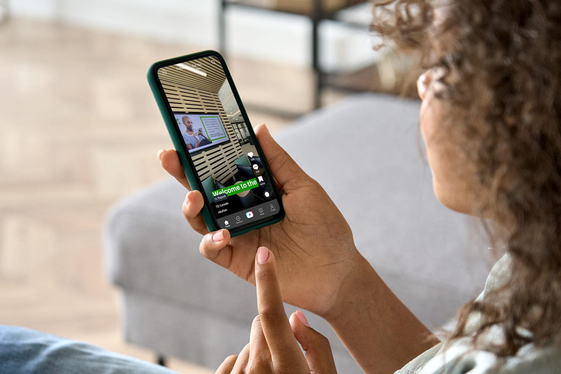 Curly-haired person lounging on a couch a watching a social media reel on their phone featuring the TD brand.
