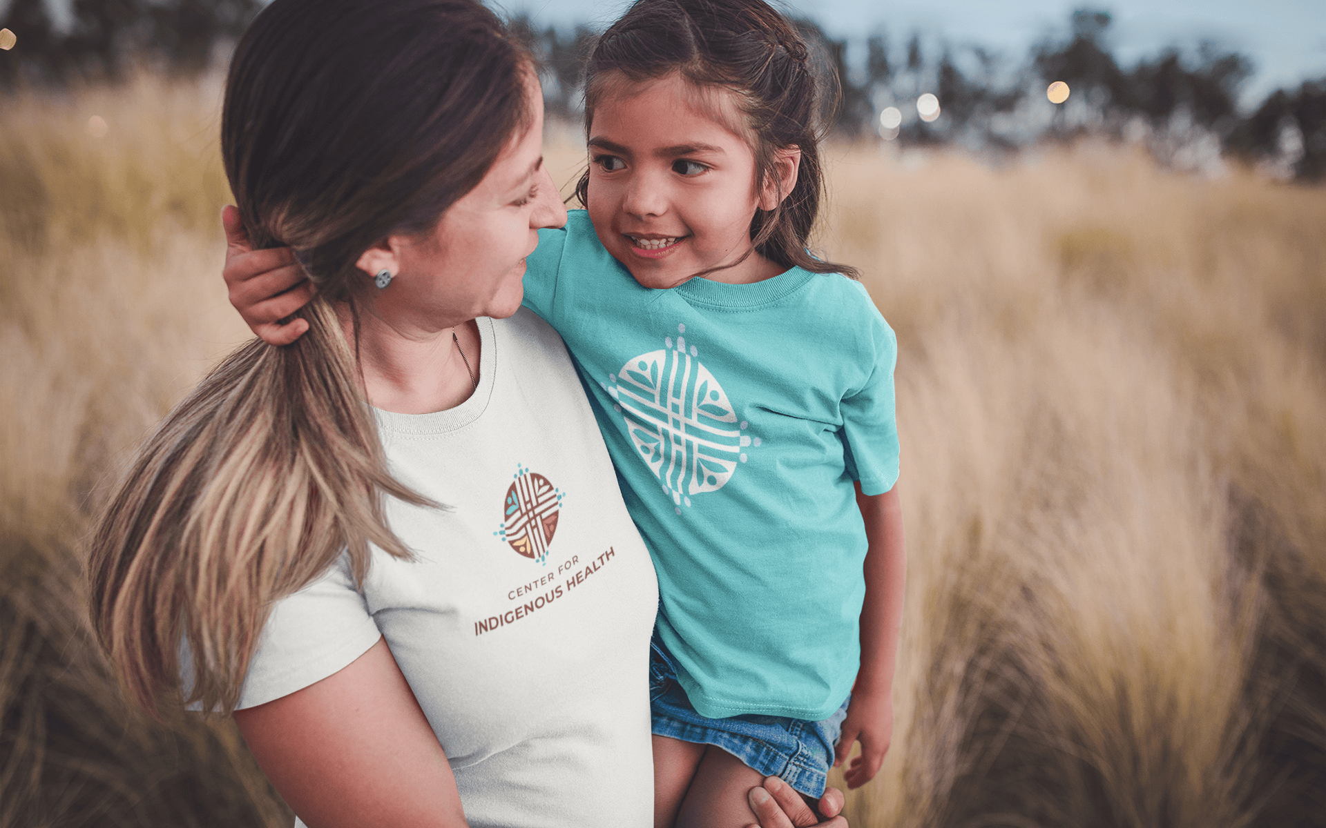 Mother holding a daughter while standing in a field, wearing t-shirts with the logo for the Center for Indigenous Health