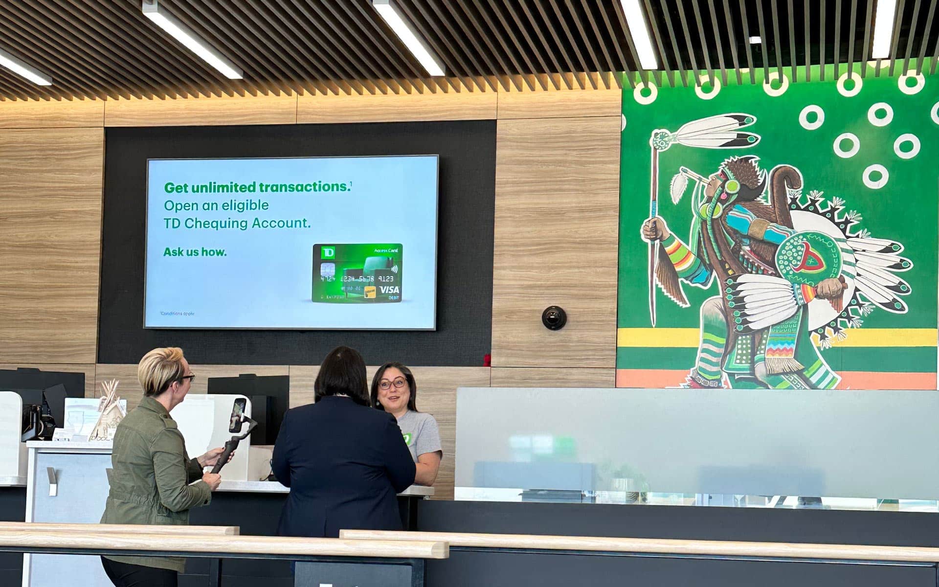 A TD Bank front desk where an advisor speak to the DDP team while filming. The desk is in front of a large mural of an Indigenous man in regalia.