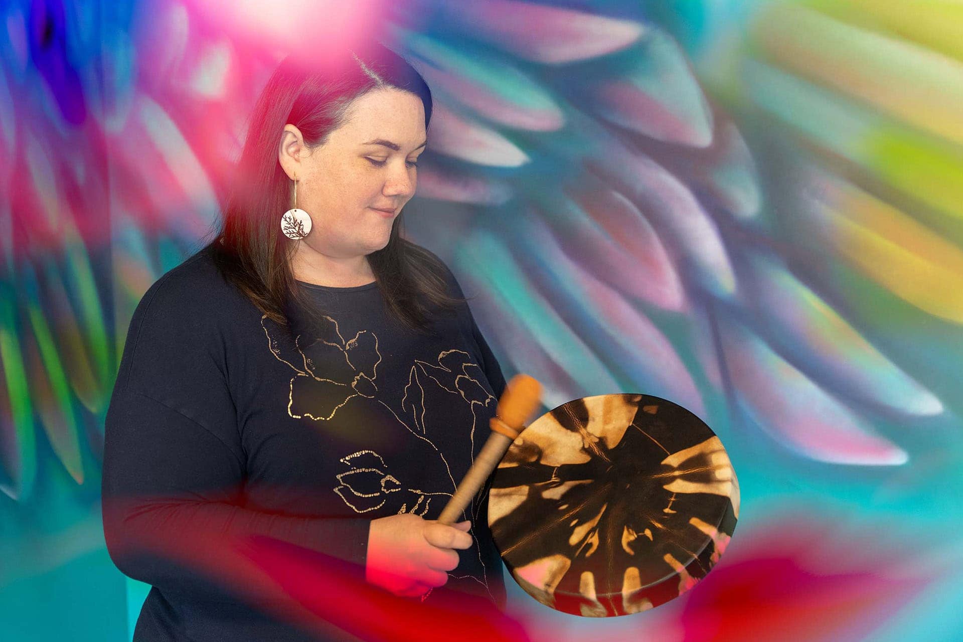 Long black hair light skinned woman with round earrings playing a hand drum in front of a mural with colourful feathers.