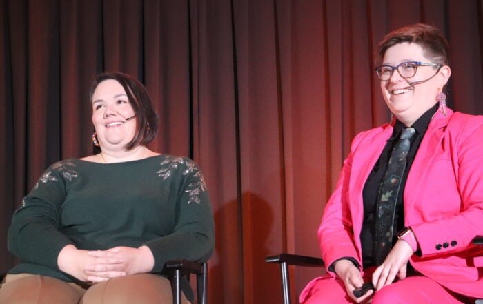 Two light skinned women sitting in director style chairs on stage with headset microphones with a dark curtain behind them.