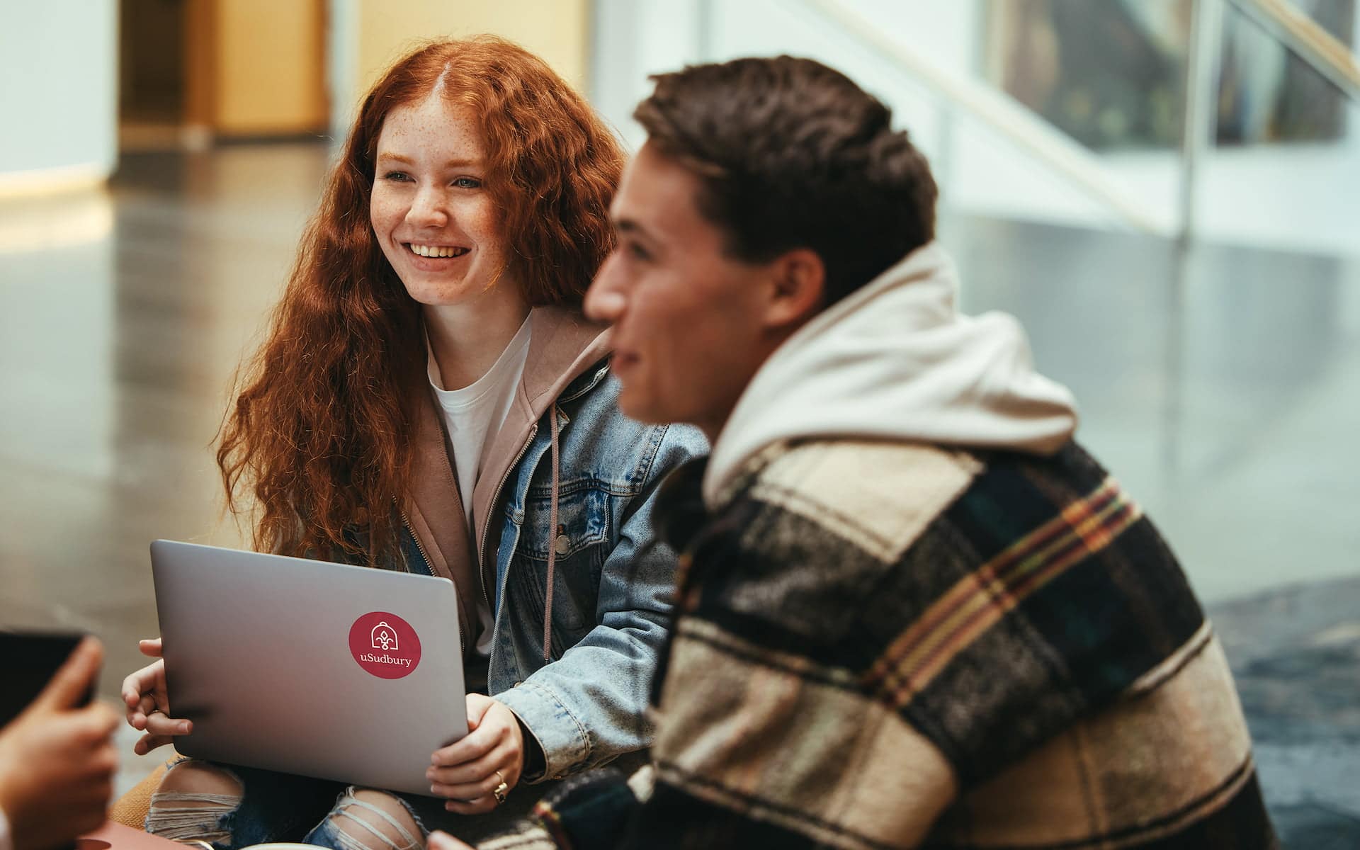 Young woman with long curly red hair sitting on the floor with fellow students, with a laptop on her lap. The laptop has a Université de Sudbury logo.