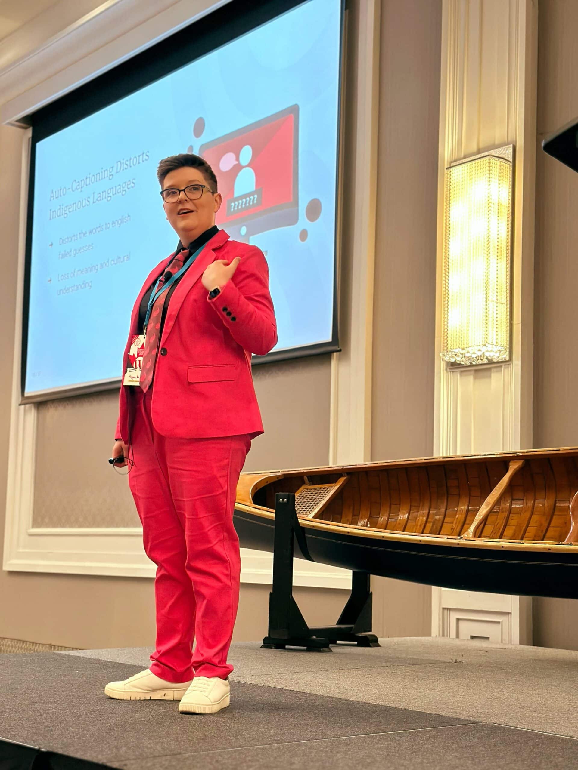 8da1817b6f63aa4322c3ce8fc4361dfe04078ea1 (1) Meggan Van Harten giving a talk on stage at a well-lit conference venue, standing in front of a projected presentation.