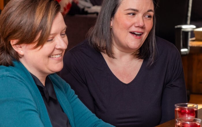 Two women smiling at the camera while sitting at an office desk, with two drinks and two balls of yarn on the desk.